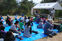 屋外の広場で参加者たちがブルーシートの上に座って説明を聞いている写真