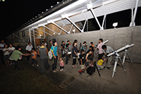 夜の屋外観測スペースで親子や参加者たちが集まって望遠鏡を囲み星空観察をしている写真