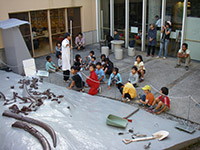 施設前の屋外スペースで子供たちが地面に並べられた化石標本を見ながら説明を聞いている写真