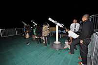 夜の屋上で参加者たちが望遠鏡を使って月や星を観察している写真