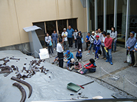 施設前の屋外スペースで親子や参加者たちが化石標本を囲んで解説を聞いている写真