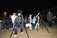 夜の広場で参加者たちが複数の望遠鏡を使って天体観測を行っている写真