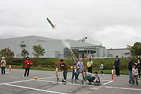 屋外の広場で親子や参加者たちが協力して実験や競技に取り組んでいる写真