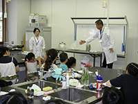 実験室で白衣を着た講師が子供たちに実験の説明をしている写真