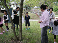 林の中で講師と子供たちが木の周りに集まり自然観察をしている写真