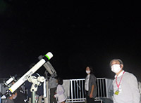 夜空の下で空を見上げる男性とその隣に望遠鏡が設置されている様子の写真