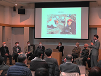 スクリーンの前で整列して座り談笑しているスーツ姿の男女の写真