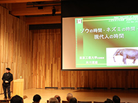 スクリーンに表示されたゾウの写真とその横に立つ黒色の服を着た1人の男性の写真