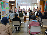 ステージ上に立ち発表を行う子供たちとそれを着席して見ている大人たちの写真