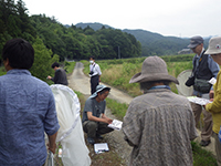 畑の横の歩道上でしゃがみ込み白い紙にスケッチをする男性