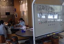 文字が書かれたホワイトボードとホワイトボードの奥で机に向かい作業をしている女性の写真