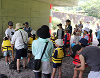 河川敷でライフジャケットを着用した児童と保護者たちが集まっている様子の写真