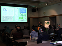 暗い照明の部屋でスクリーンに映し出された資料を見ている参加者の後ろ姿の写真