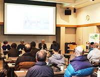 照明の明るい部屋でスクリーンの真下でマイクを持ち話をする男性とその話を聞く参加者たちの後ろ姿の写真