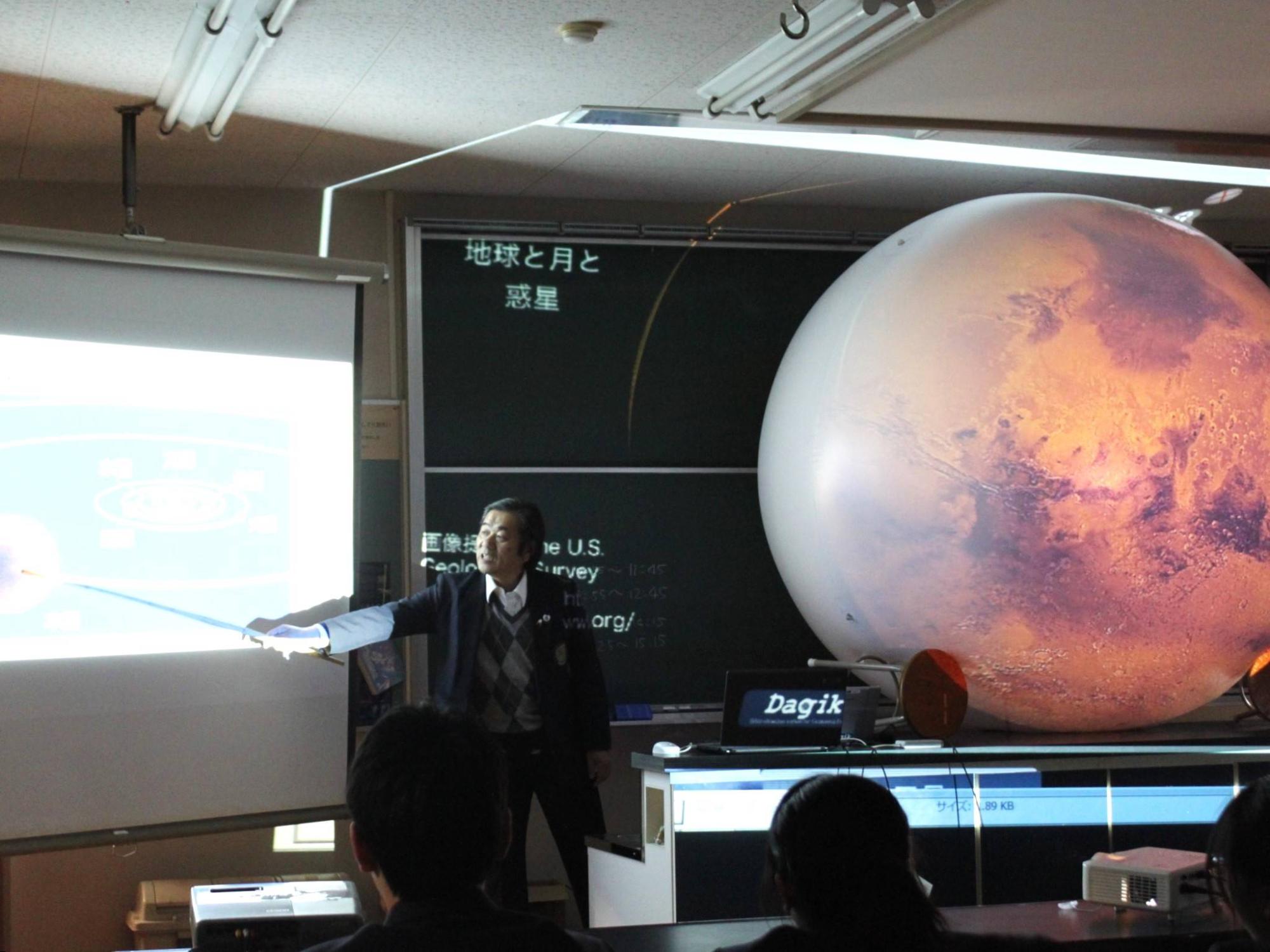 球体投影装置の隣で惑星について説明をしているスーツ姿の男性の写真