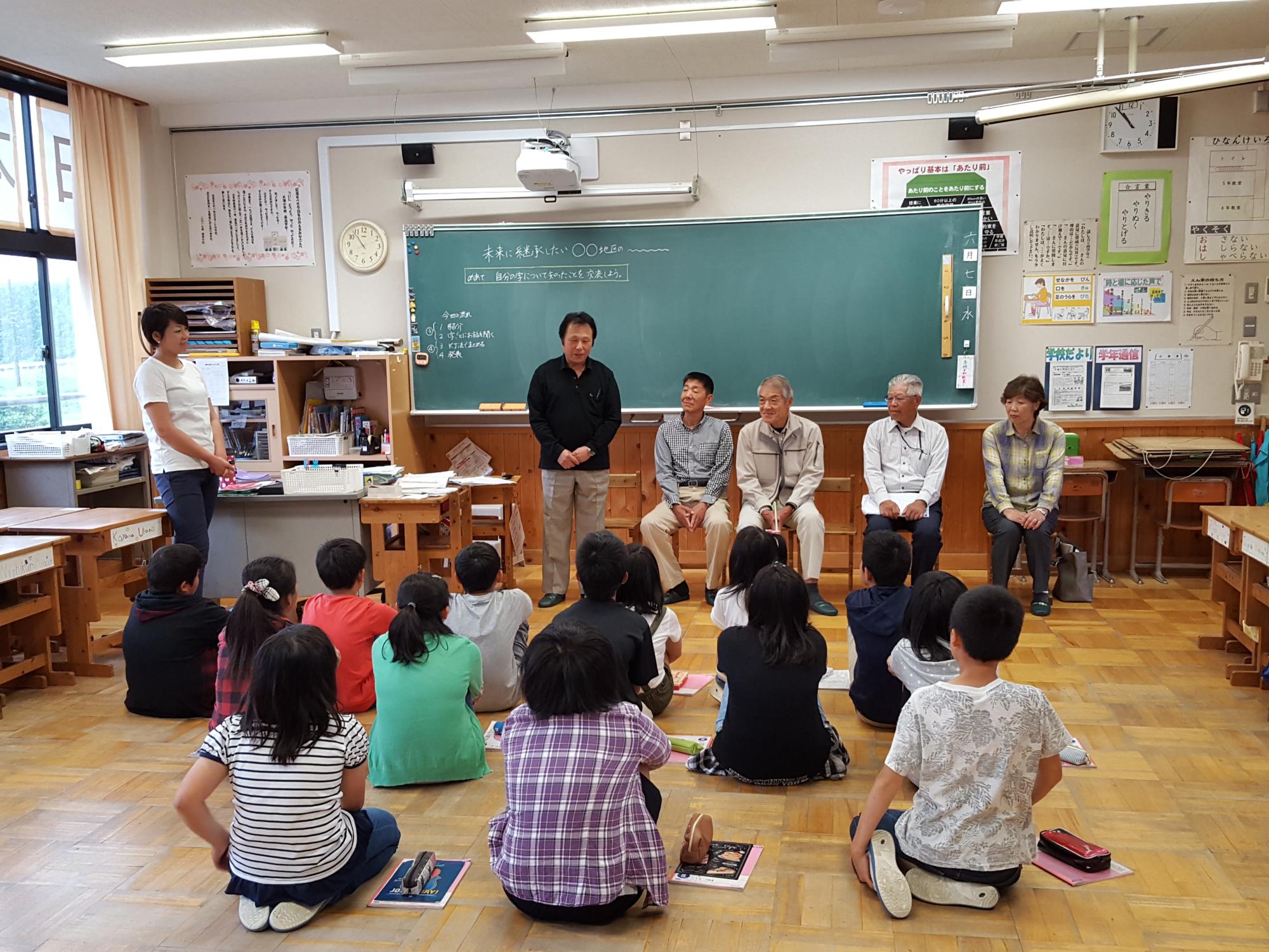 並んだ椅子に座っている大人4人と立って話している大人の話を床に座って聞いている児童たちの写真