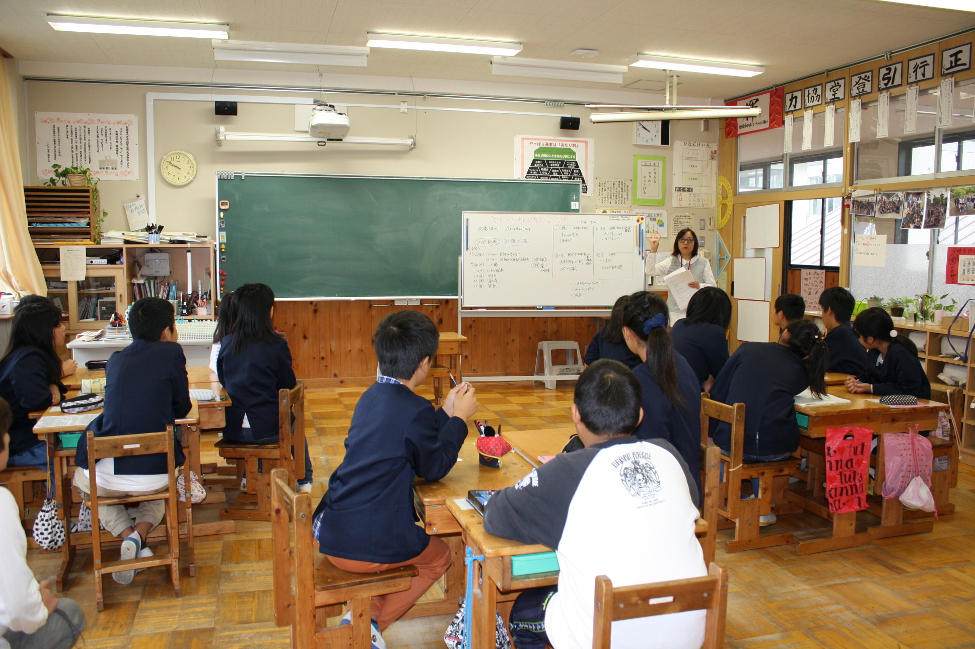 ホワイトボードに書かれた資料を基に話す先生の話を聞く児童たちの写真