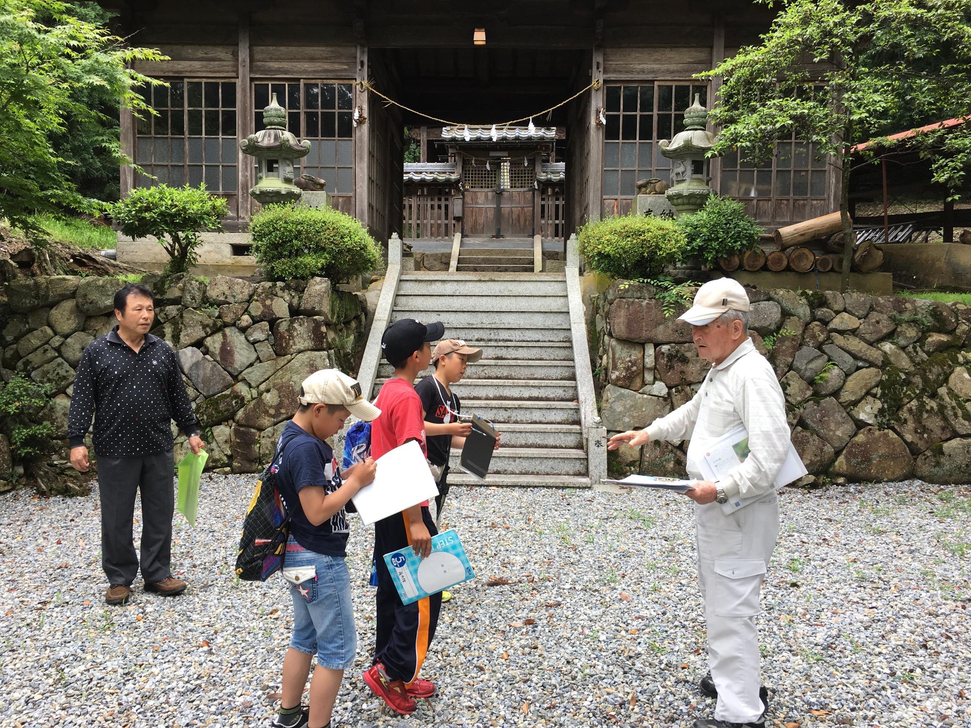 神社の敷地内で男性の話を聞いてメモを取る児童たちの写真