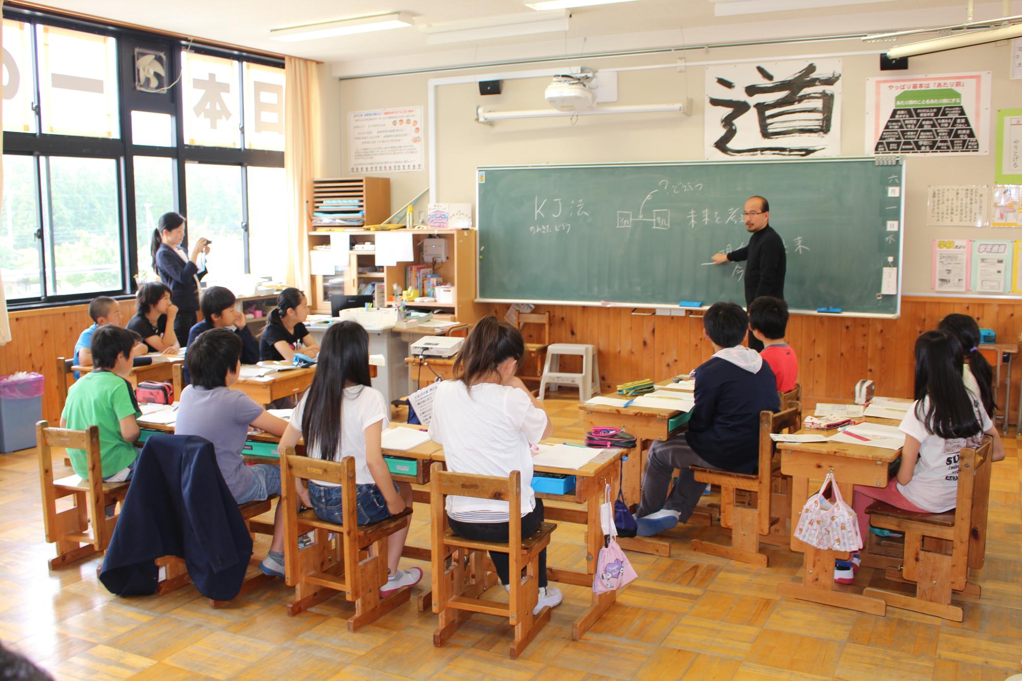 コの字型に並んだ席に座って先生の話を聞く児童たちの写真