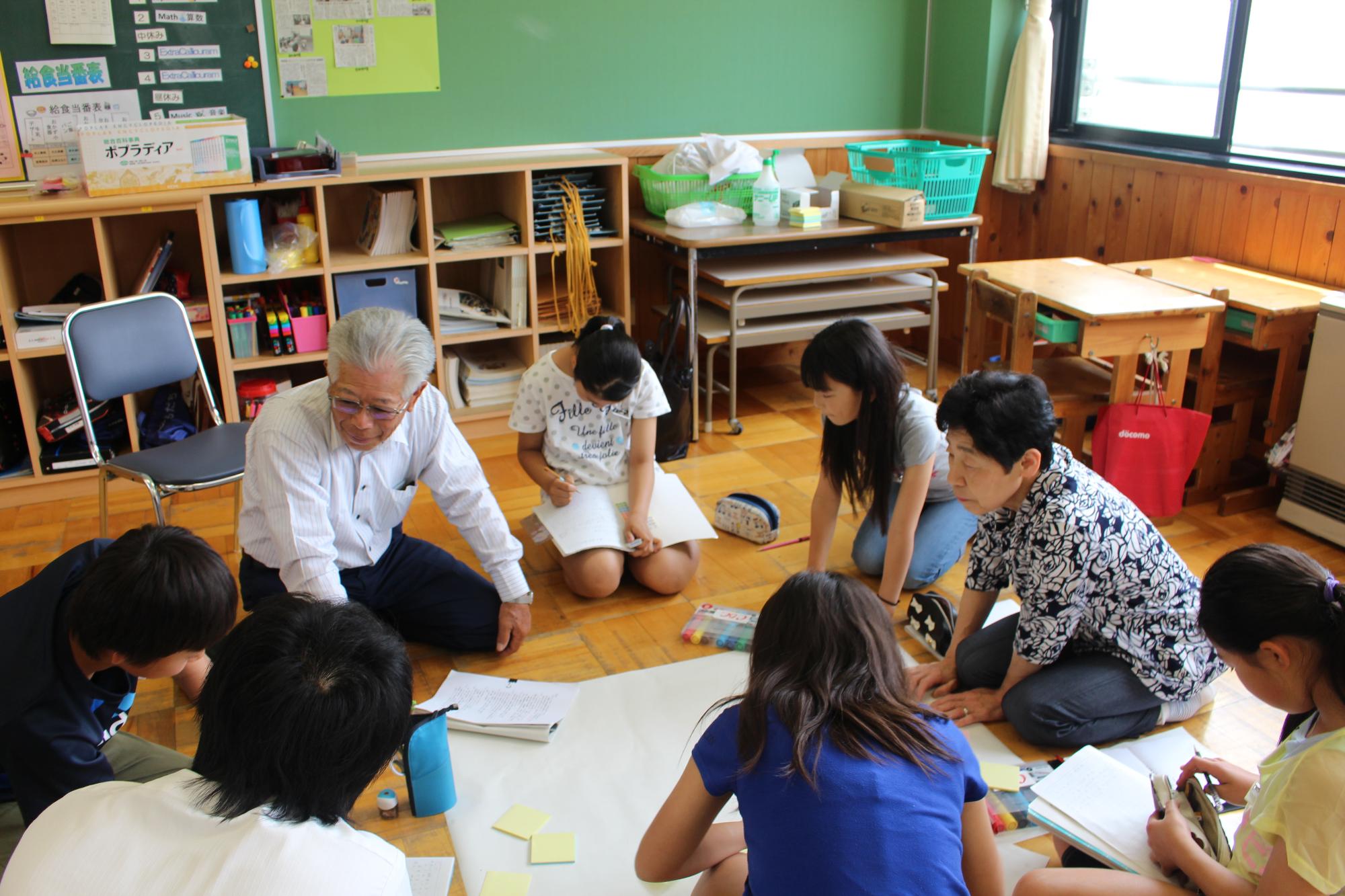 教室の床に広げた模造紙を取り囲んで座っている児童たちと大人2人の写真