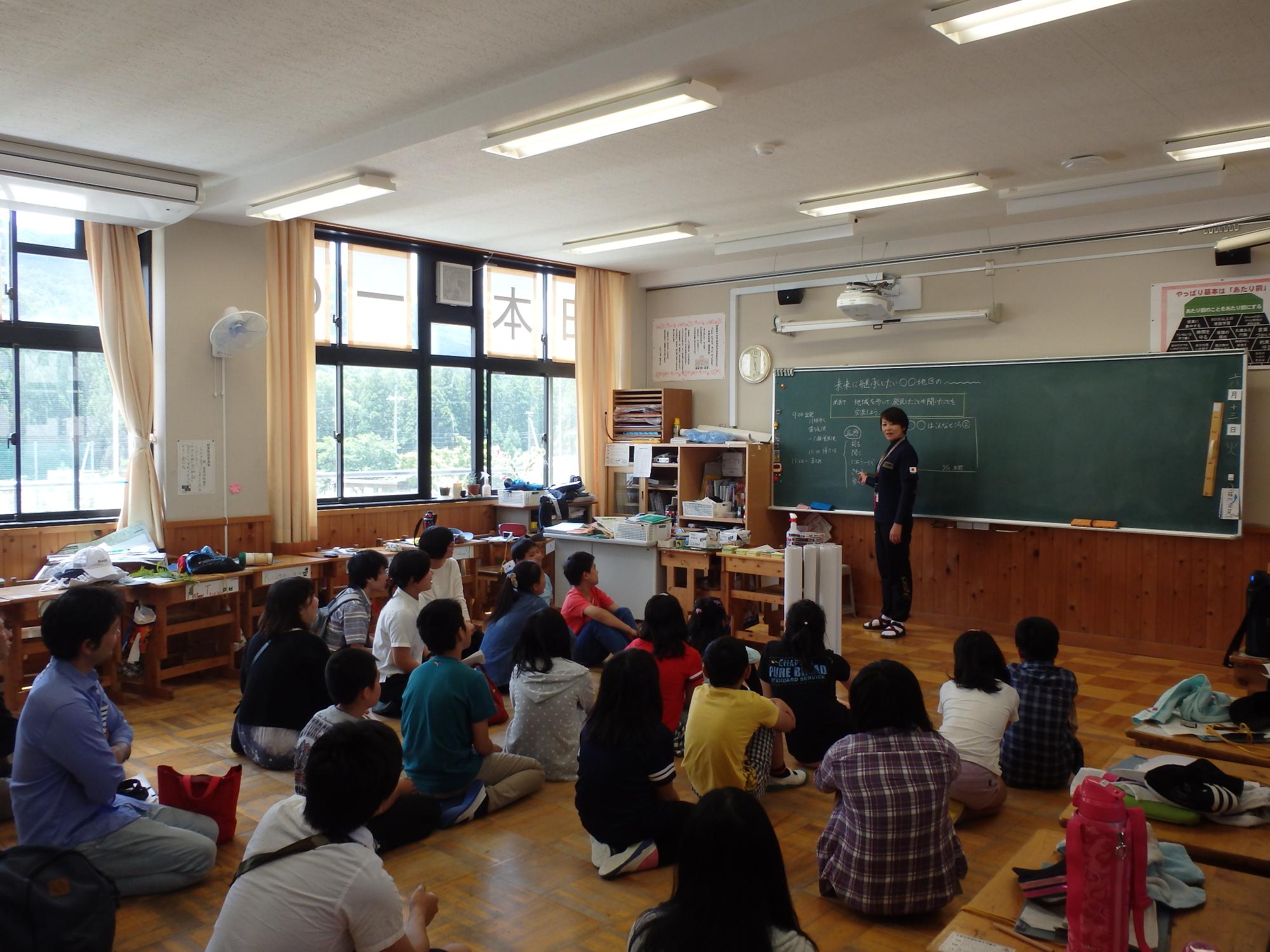 黒板に書いた資料を基に話す先生の話を教室の床に座って聞く児童たちの写真