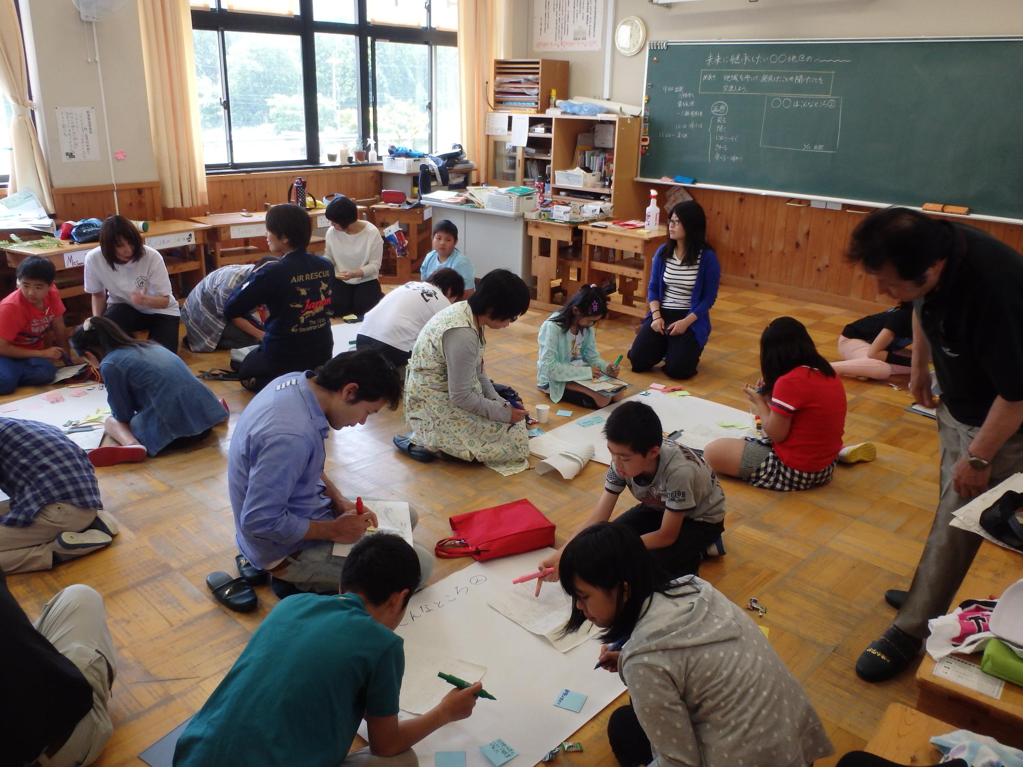 教室の床でグループごとに模造紙を広げて文字を書き込む児童たちと見守る大人たちの写真