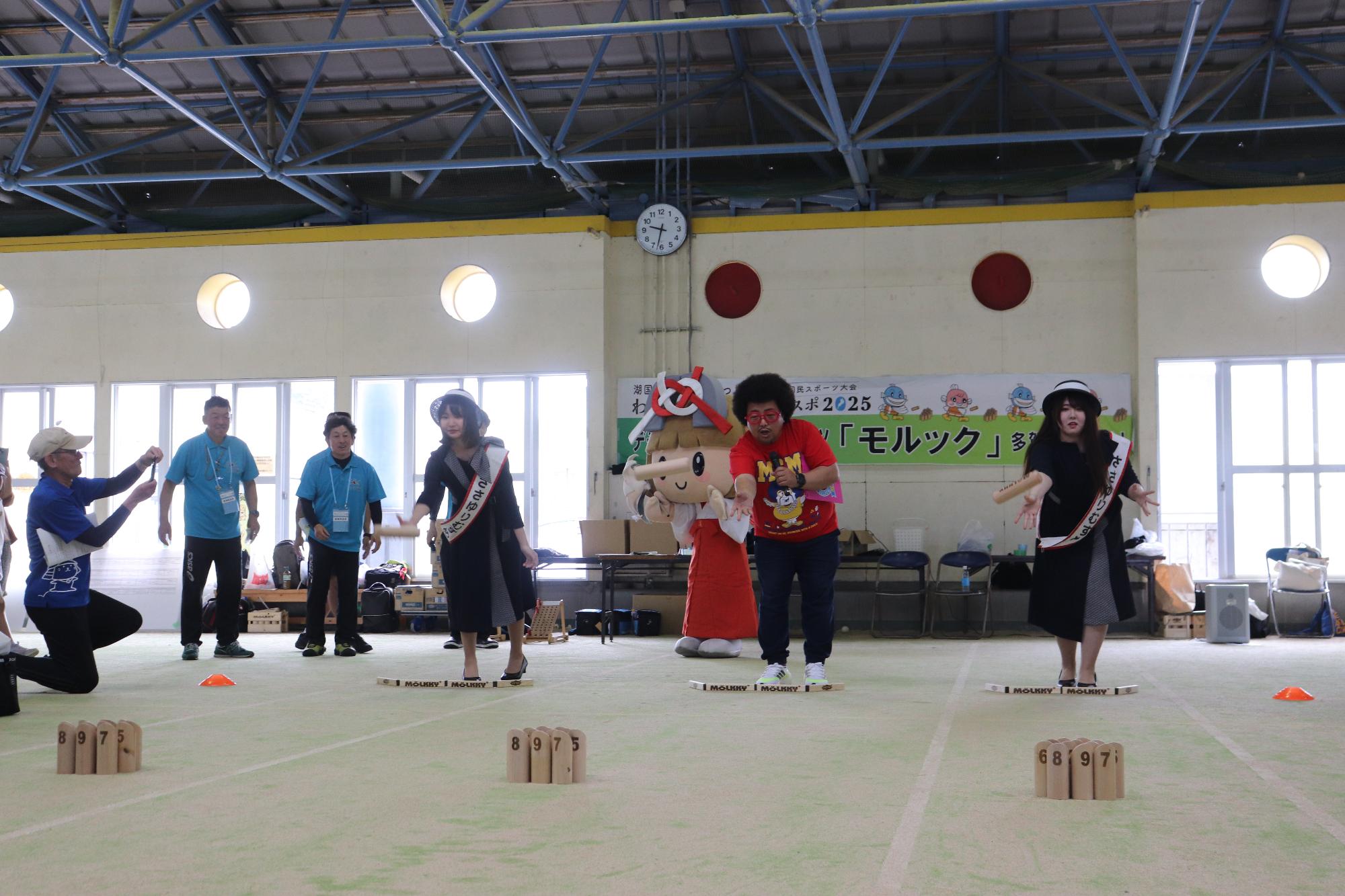 屋内会場の競技エリアでMC MORIYAさん、ささゆりむすめさんとキャラクターがモルックの始投式をしている写真