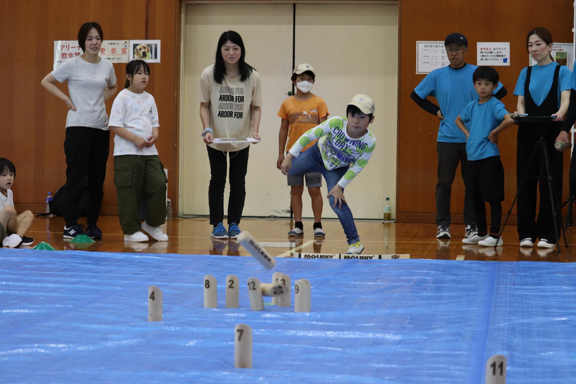 屋内会場で子どもがモルックを投げ家族やスタッフが見守っている写真