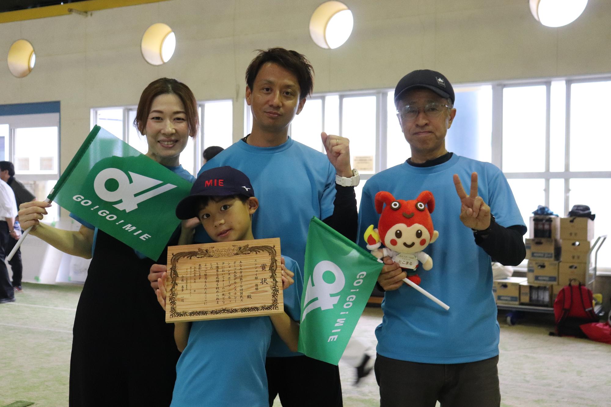 屋内会場で旗と表彰状を手にしたパーシモン津のチームメンバーが記念撮影している写真