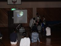 体育館で参加者たちが床に座りスクリーンに映された映像を見ながら解説を聞いている写真