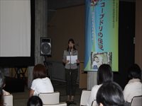 ホールの演台で講師がマイクを使って来場者に向けて講演を行っている写真