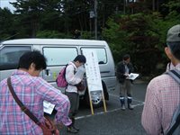 駐車場で参加者たちが掲示された案内板を見ながら説明を聞いている写真