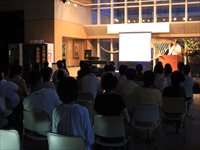 夜のホールで参加者たちが椅子に座りステージとスクリーンを見ながら公演を鑑賞している写真