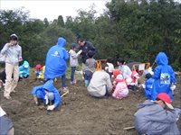 屋外の広場で親子や参加者たちが地面に座って野外活動の説明を聞いている写真
