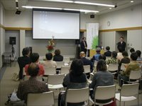 会議室の前方で講師が参加者たちに向けて講演を行っている写真