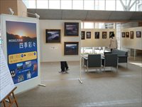 大型のパネルに写真展のポスターが貼られその奥で様々な写真が飾られている展示風景の写真