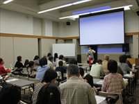 会議室で講師がスクリーンを使って参加者たちに講義を行っている写真