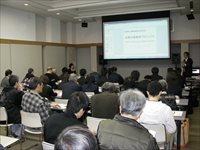 会議室で参加者たちが椅子に座りスクリーンを見ながら研究発表を聞いている写真
