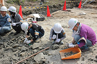 ヘルメットをした子供と大人が地面にしゃがんで土を見ている写真