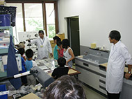 子供たちと白衣を着た男性が実験室で輪になって話している写真