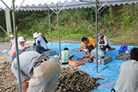 ブルーシートの上に土が積まれ、しゃがんで作業する子供と大人たちの写真