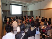 前方のスクリーンを見て話を聞く椅子に座った男女を後ろから撮影した写真