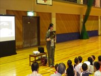 体育館の舞台前で講師がマイクを持って児童たちに向けて話をしている写真