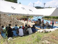 屋外のテントの下で参加者たちが地面に座って説明を聞いている写真