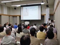 会議室で大勢の参加者がスクリーンを見ながら発表を聞いている写真