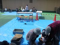 体育館の床で帽子をかぶった参加者たちがしゃがんで資料や道具を確認している写真