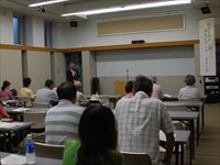 会議室で参加者たちが机に着席して前方の説明を聞いている写真