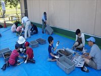 屋外のシートを敷いた作業スペースで親子や児童たちが道具を使って作業をしている写真