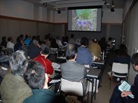 会議室で参加者たちが着席して前方のスクリーンに映し出された発表資料を見ながら説明を聞いている写真