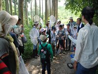 森の中で子供と大人が昆虫観察の説明を受けている写真